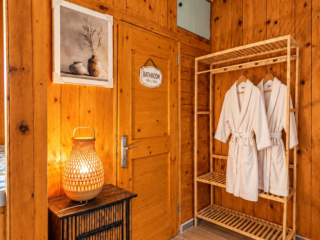 « Le Chalet » – Mountain Retreat: wood-panelled entry with a glowing bamboo lantern and two white bathrobes on a wooden rack beside the bathroom door